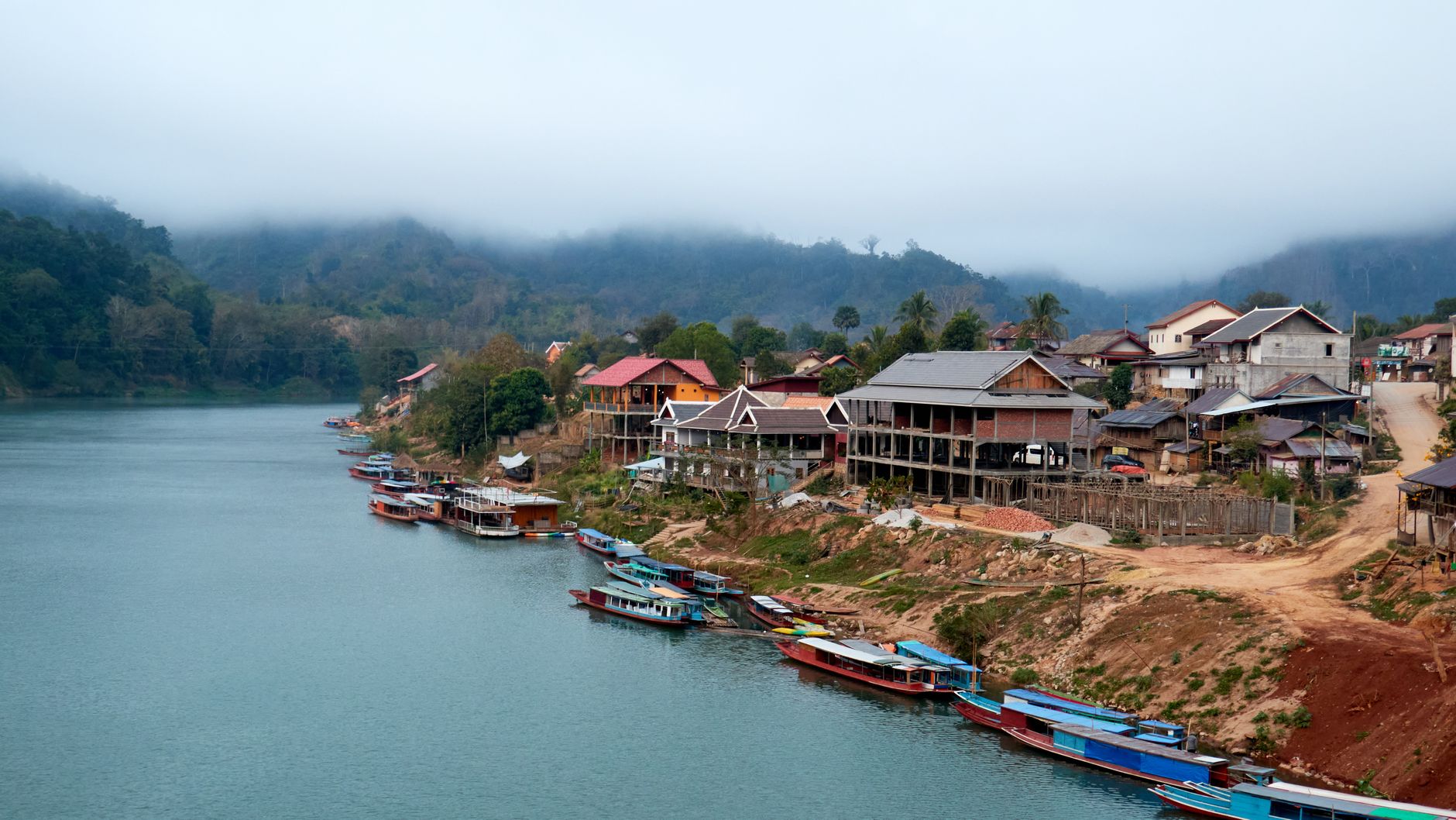 Lunghe barche colorate ormeggiate presso il villaggio nebbioso sulle rive del fiume di Nong Khiaw, Laoscon montagne ricoperte di giungla che si ergono sullo sfondo