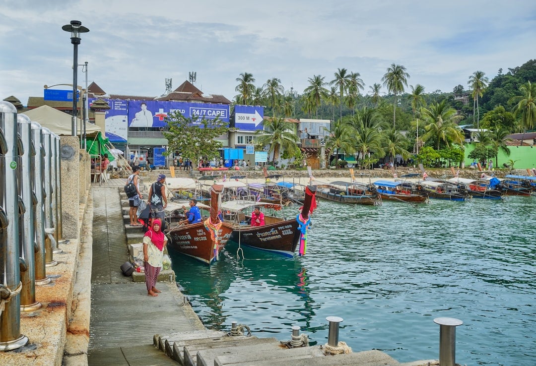 Barche a coda lunga ormeggiate al molo thailandese, con palme e costa tropicale sullo sfondo.