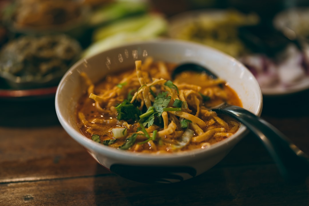 Semangkuk Khao Soi, sup mie kari santan khas Thailand Utara, dihiasi dengan rempah segar dan mie renyah di atas meja kayu.