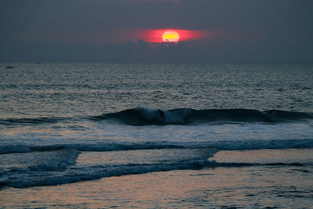 Surfers riding waves at sunset on a Bali beach, with a glowing red sun low on the horizon over the Indian Ocean