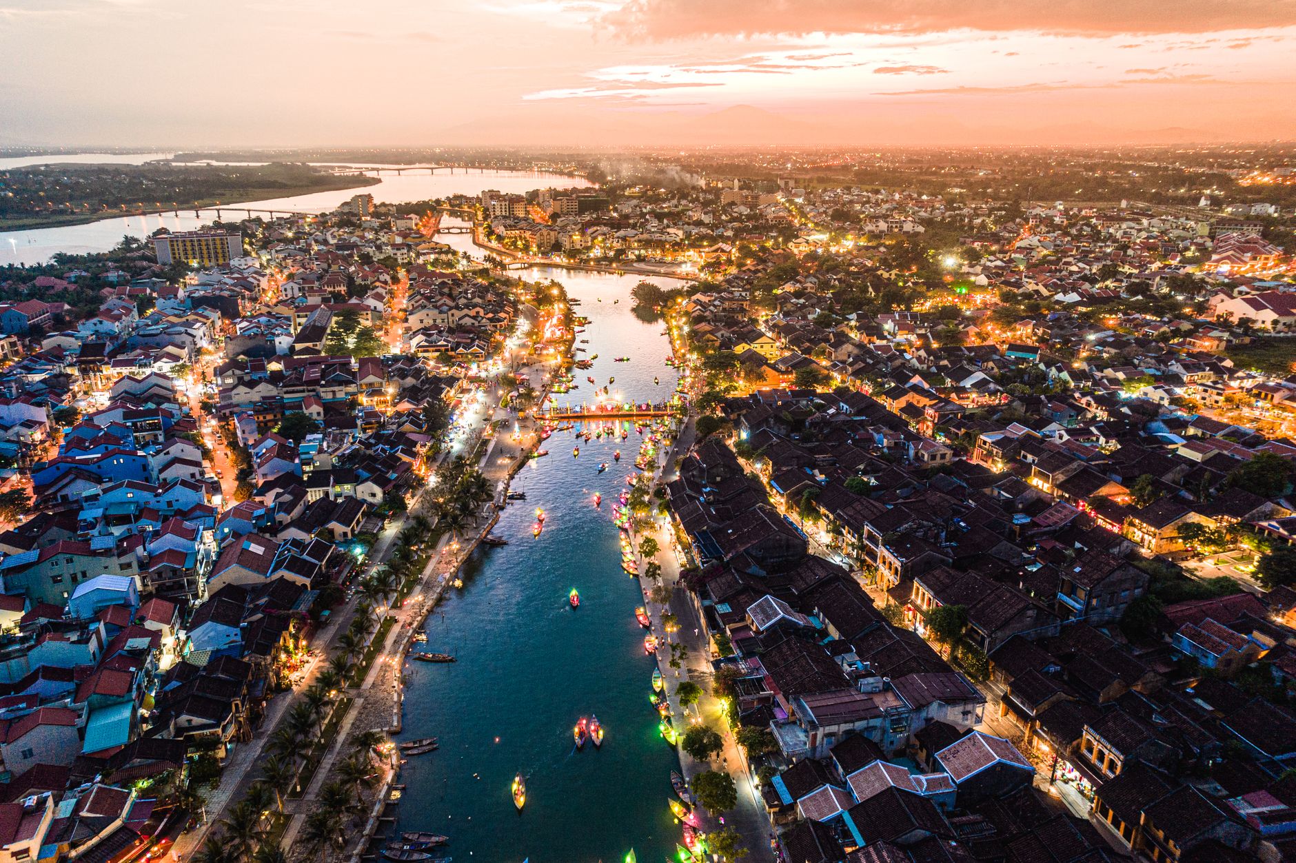 Aerial view of Hoi An ancient town at sunset with the Thu Bon River glowing with lantern boats and city lights reflecting on the water
