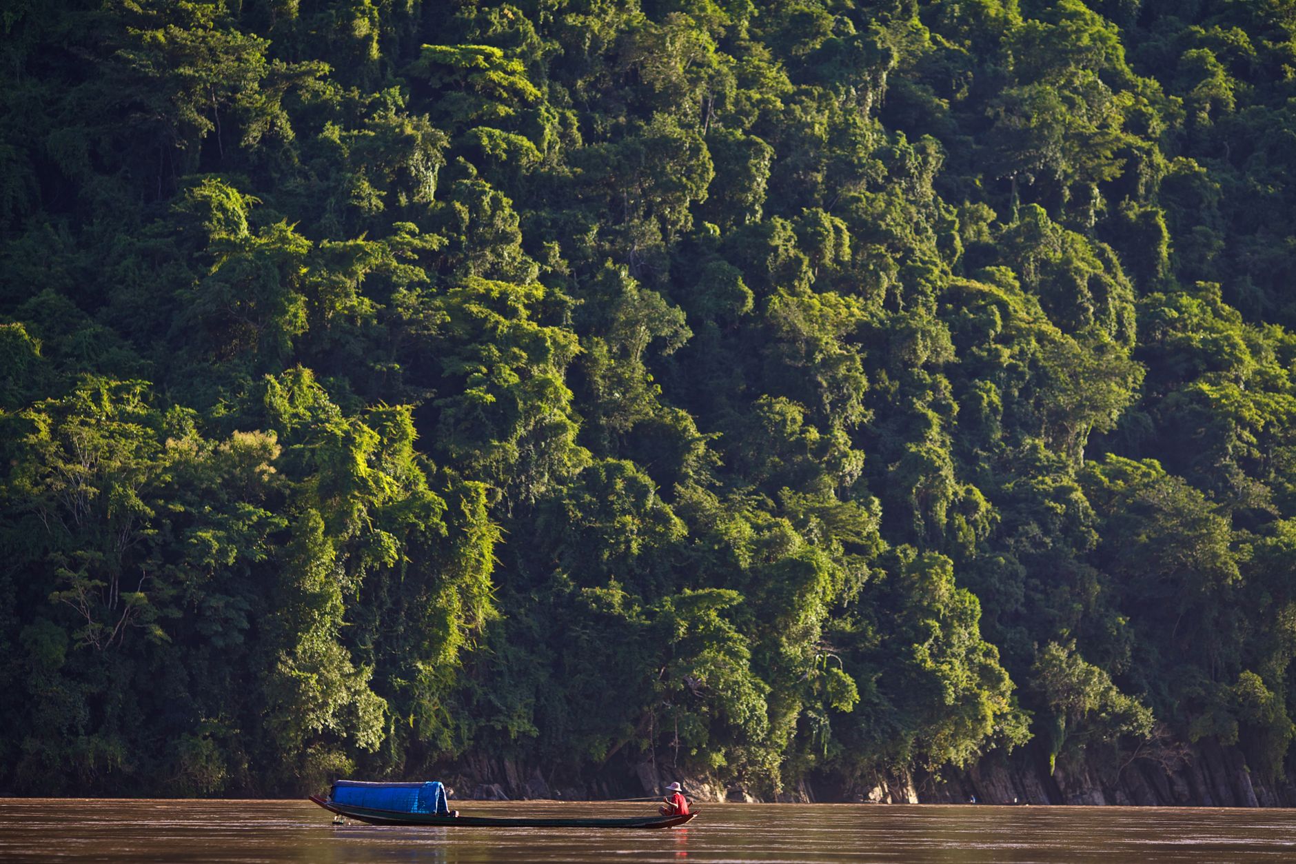 A small wooden boat traveling along the Mekong River with dense green jungle hillside in the background, Laos