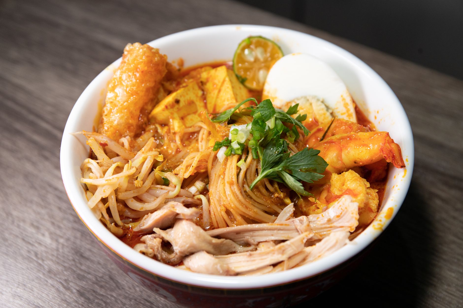 Bowl of Sarawak-style laksa noodle soup with shredded chicken, prawns, bean sprouts, egg, and a rich spiced coconut broth.