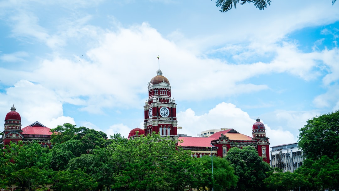 The Yangon High Court building with its iconic red-brick clock tower rising above the surrounding trees, under a blue sky with clouds.