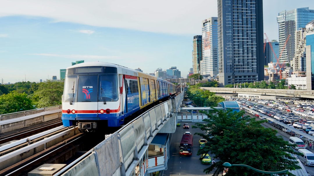 Bangkok BTS Skytrain on elevated tracks above gridlocked city traffic, with modern skyscrapers and green trees visible alongside the rail line