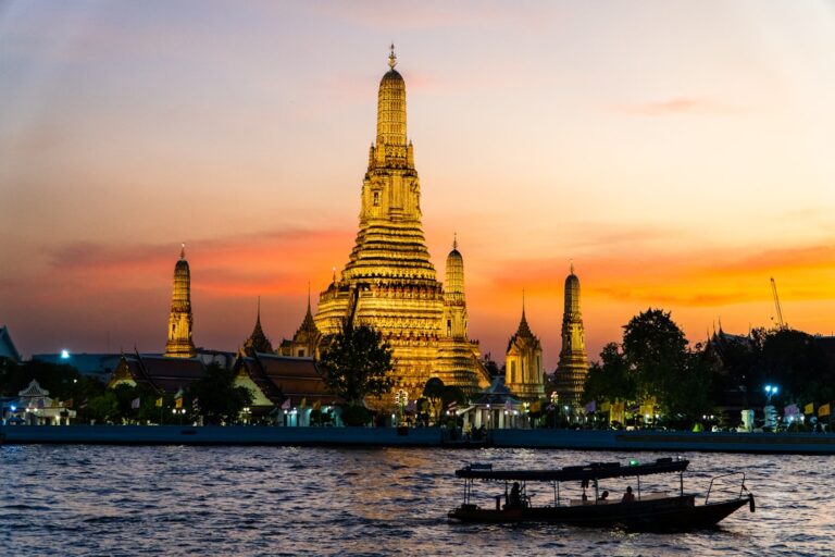 Wat Arun temple illuminated at sunset on the Chao Phraya River in Bangkok, Thailand, with a longtail boat silhouette in the foreground