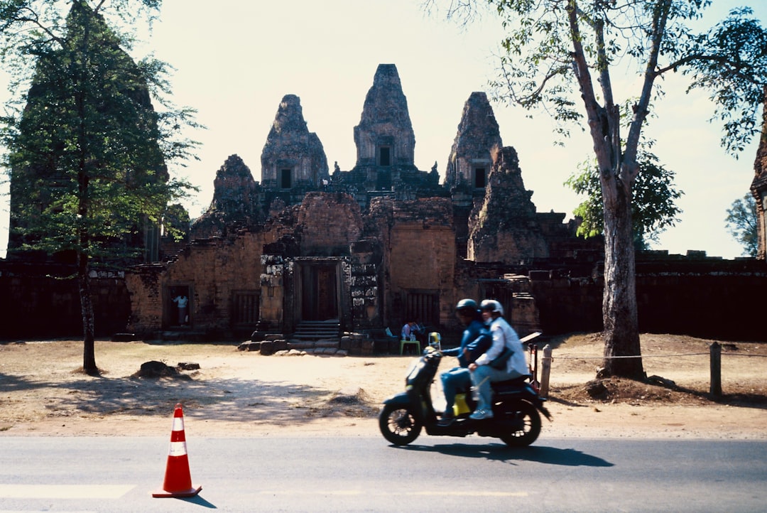 Two people riding a scooter past ancient temple ruins in Southeast Asia