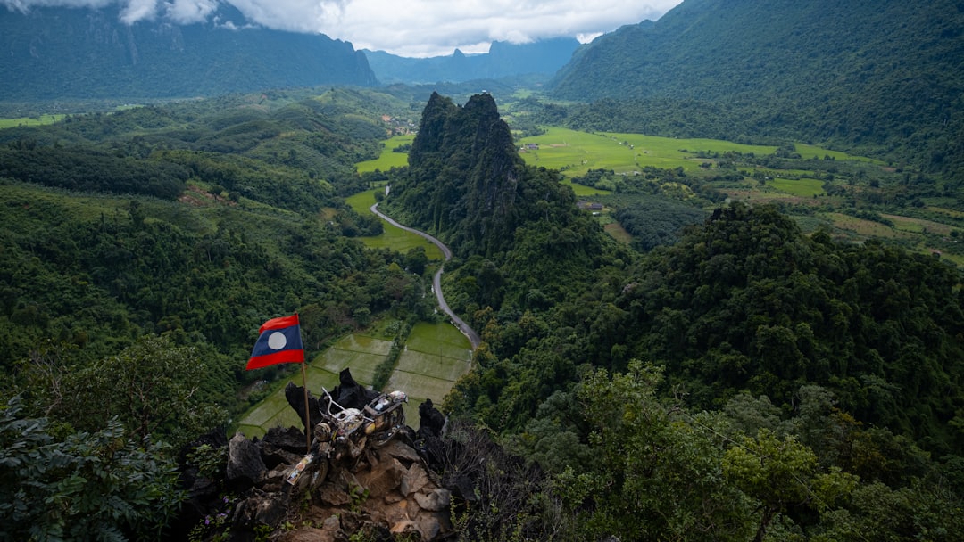 Panoramic view from Nam Xay viewpoint near Vang Vieng, Laos, showing karst peaks and valley below with Lao flag in foreground