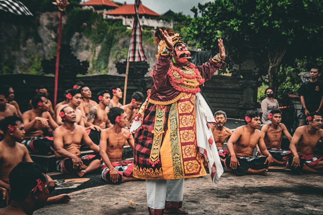 A Kecak performer in an elaborate ornate mask and traditional Balinese costume dances before a seated shirtless male chorus during a Kecak fire dance performance in Bali
