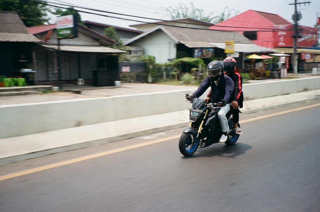 A Honda MSX motorcycle with a rider and passenger moving through a Thai town street, with traditional wooden shophouses lining the road